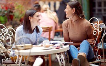 Two women sit at a round, white metal table outdoors, engaged in a lively conversation. One woman wears a light blue striped shirt, the other a beige top. Books and a drink are on the table. The background is a cozy café with plants and other customers seated.