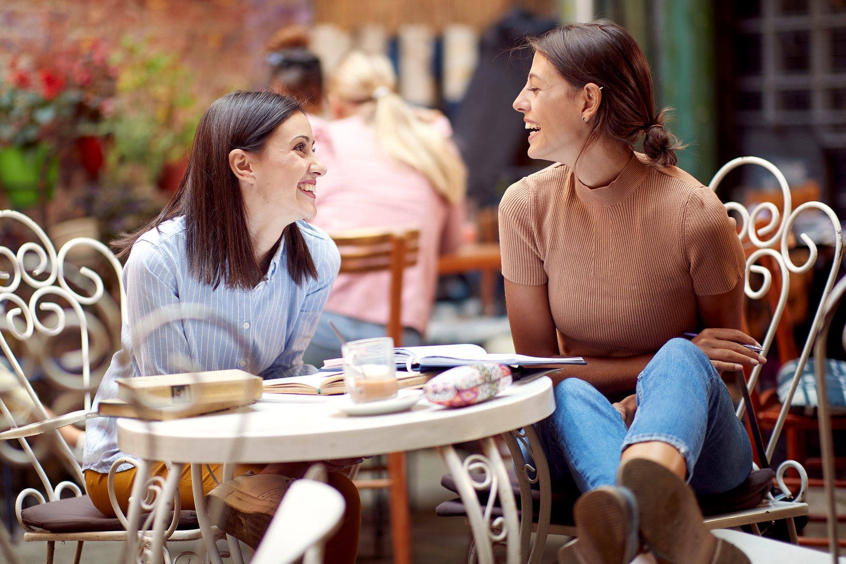 Two women sit at a round, white metal table outdoors, engaged in a lively conversation. One woman wears a light blue striped shirt, the other a beige top. Books and a drink are on the table. The background is a cozy café with plants and other customers seated.