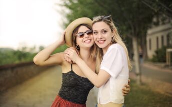 Two women stand outdoors on a pathway lined with trees. One wears a hat, sunglasses, a dark tank top, and red skirt, while the other wears a white blouse and blue jeans. They smile and embrace each other in a friendly pose. The background is blurred and green.