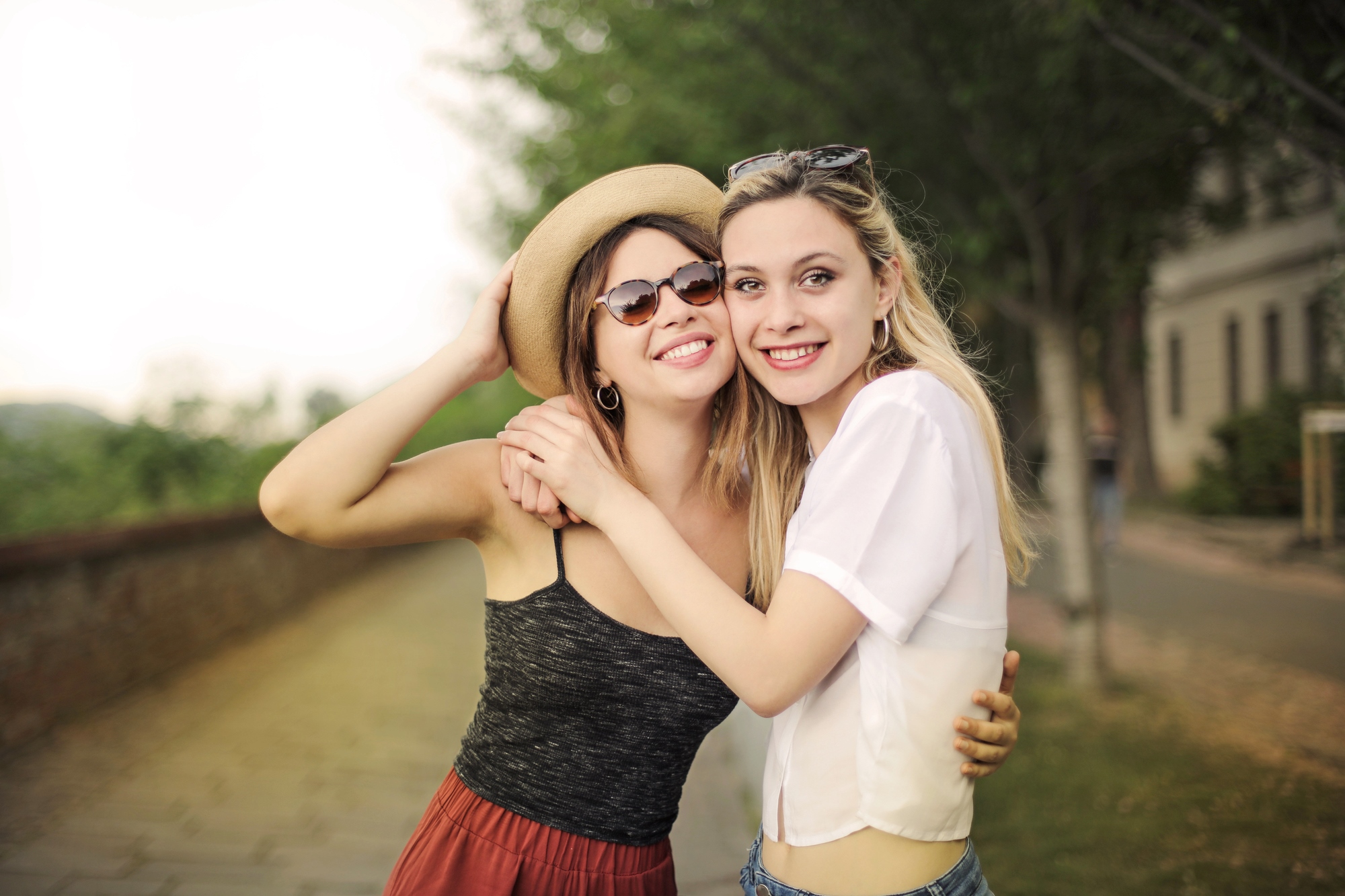 Two women stand outdoors on a pathway lined with trees. One wears a hat, sunglasses, a dark tank top, and red skirt, while the other wears a white blouse and blue jeans. They smile and embrace each other in a friendly pose. The background is blurred and green.