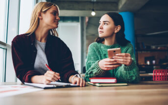 Two young women are sitting at a wooden table. One woman, holding a pen, writes on a notebook while looking at the other woman, who is wearing a green sweater and holding a smartphone, also gazing back attentively. They are in a modern, industrial-style room.