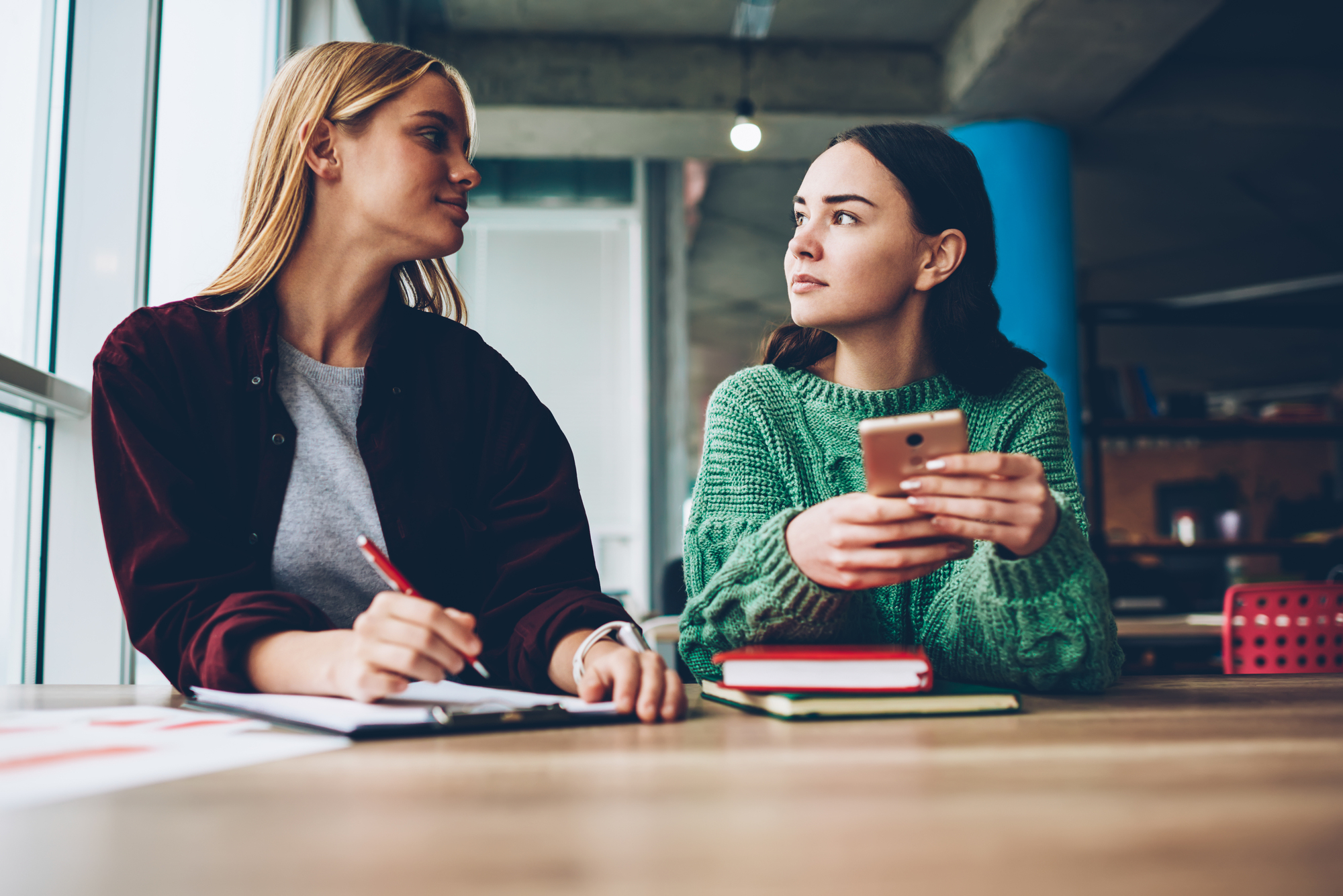 Two young women are sitting at a wooden table. One woman, holding a pen, writes on a notebook while looking at the other woman, who is wearing a green sweater and holding a smartphone, also gazing back attentively. They are in a modern, industrial-style room.