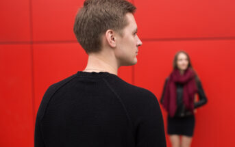 A man in a black sweater is seen from behind, looking towards a woman in the background who is wearing a black dress and a burgundy scarf. They are standing in front of a red wall.