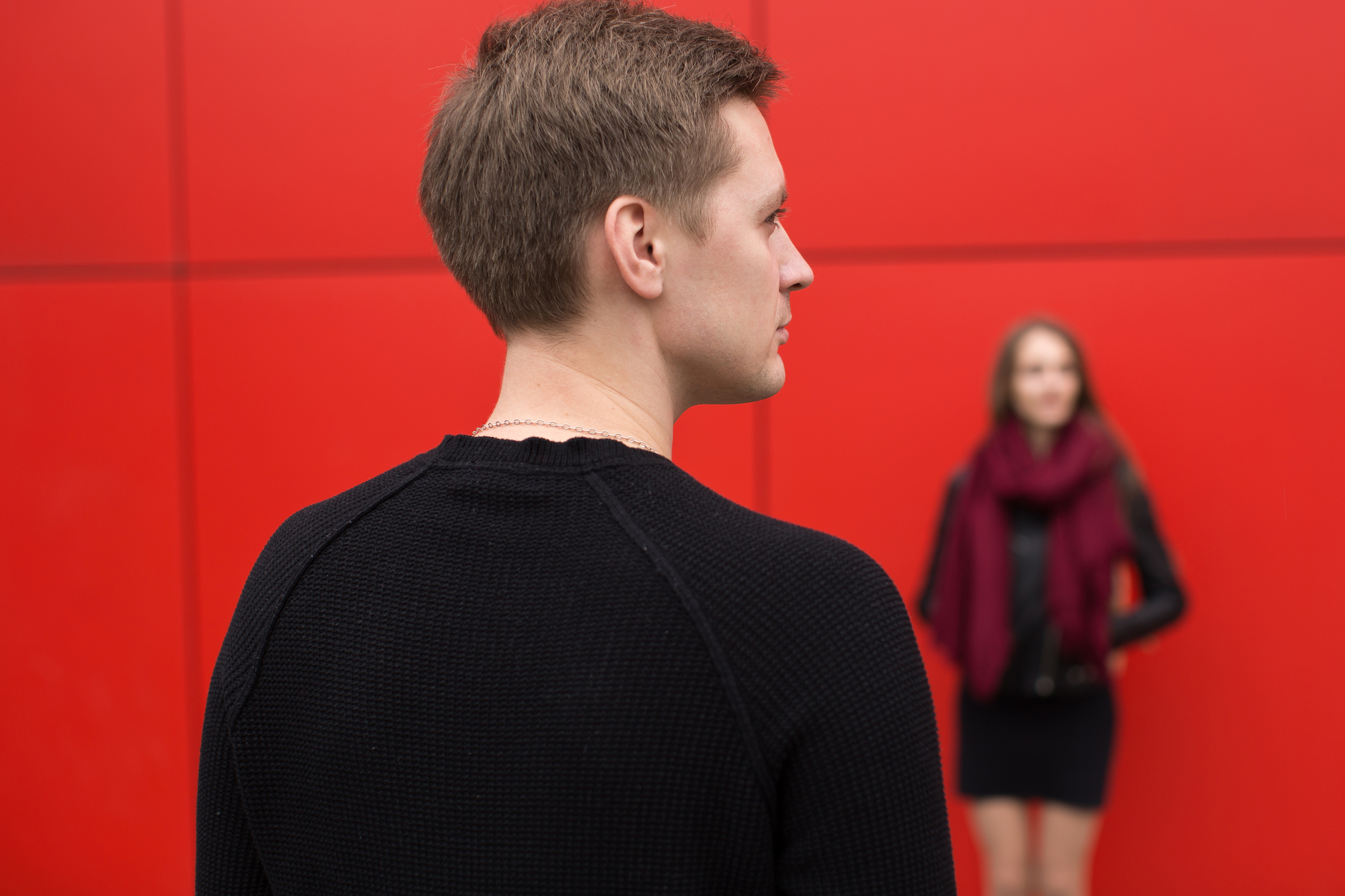 A man in a black sweater is seen from behind, looking towards a woman in the background who is wearing a black dress and a burgundy scarf. They are standing in front of a red wall.