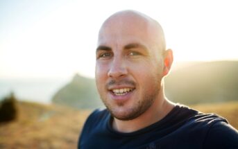 A bald man with a short beard smiles at the camera in an outdoor setting. The background shows a sunlit landscape with distant hills and a clear sky.