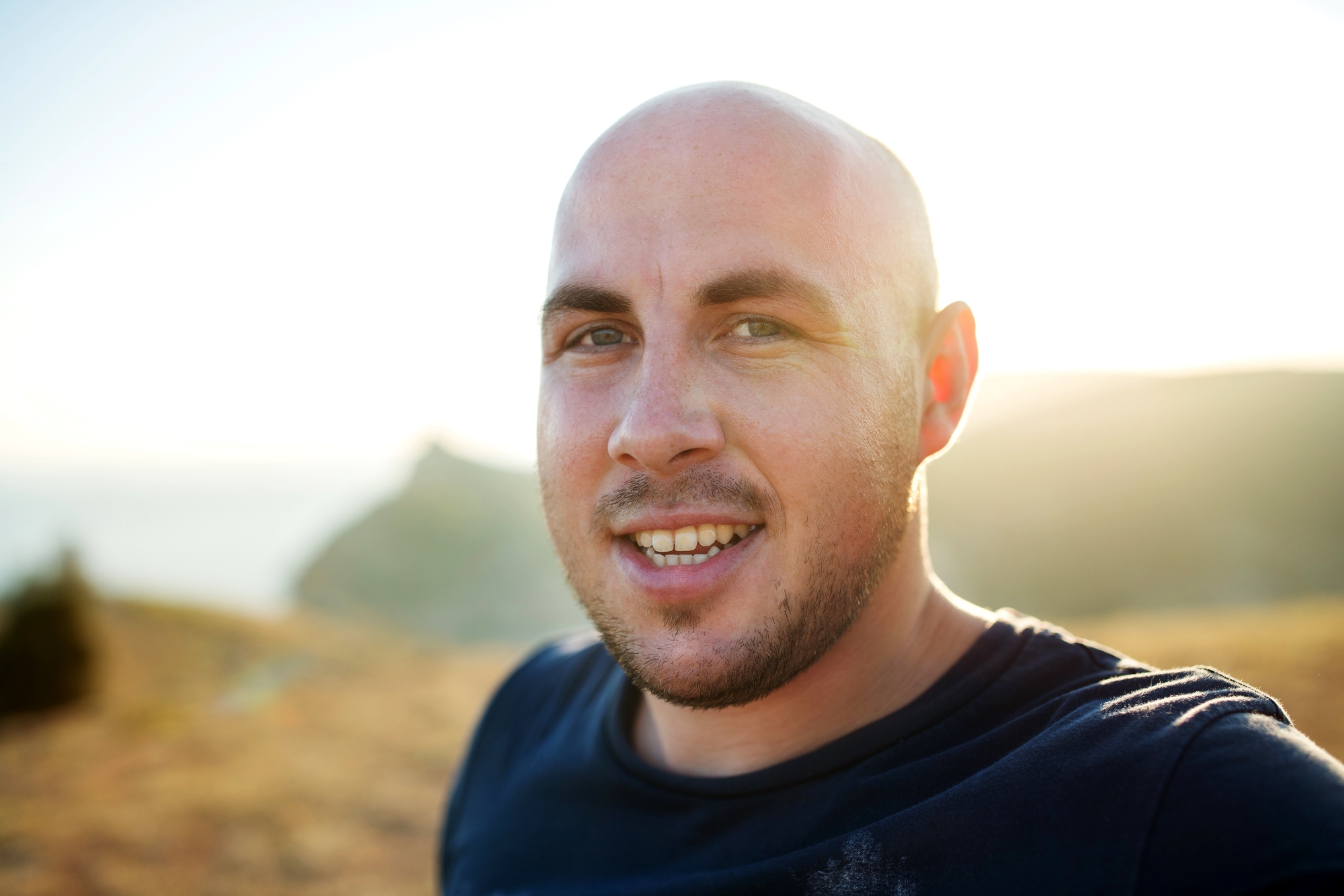 A bald man with a short beard smiles at the camera in an outdoor setting. The background shows a sunlit landscape with distant hills and a clear sky.