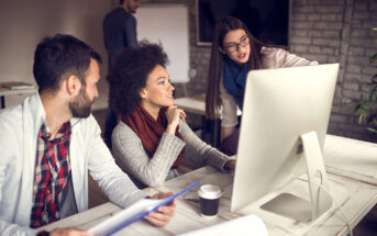 Three colleagues are gathered around a desk, engaged in discussion while looking at a computer screen. The woman standing points at the screen, while the seated man and woman listen attentively. Another person is in the background, facing a whiteboard.