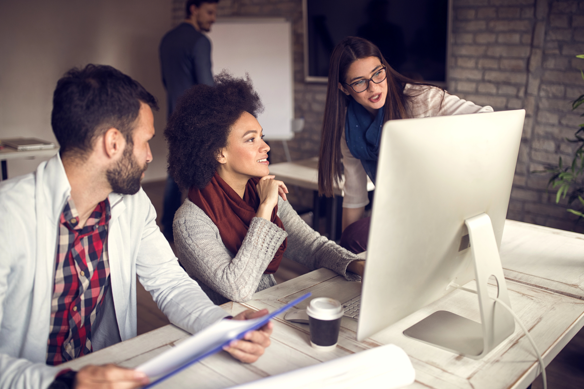 Three colleagues are gathered around a desk, engaged in discussion while looking at a computer screen. The woman standing points at the screen, while the seated man and woman listen attentively. Another person is in the background, facing a whiteboard.