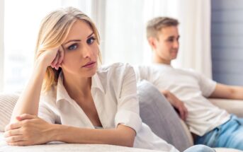 A woman with blonde hair, wearing a white shirt, sits on a couch with her hand propping up her head, looking contemplative. A man with short hair, dressed in a white t-shirt, sits slightly out of focus in the background, also on the couch.