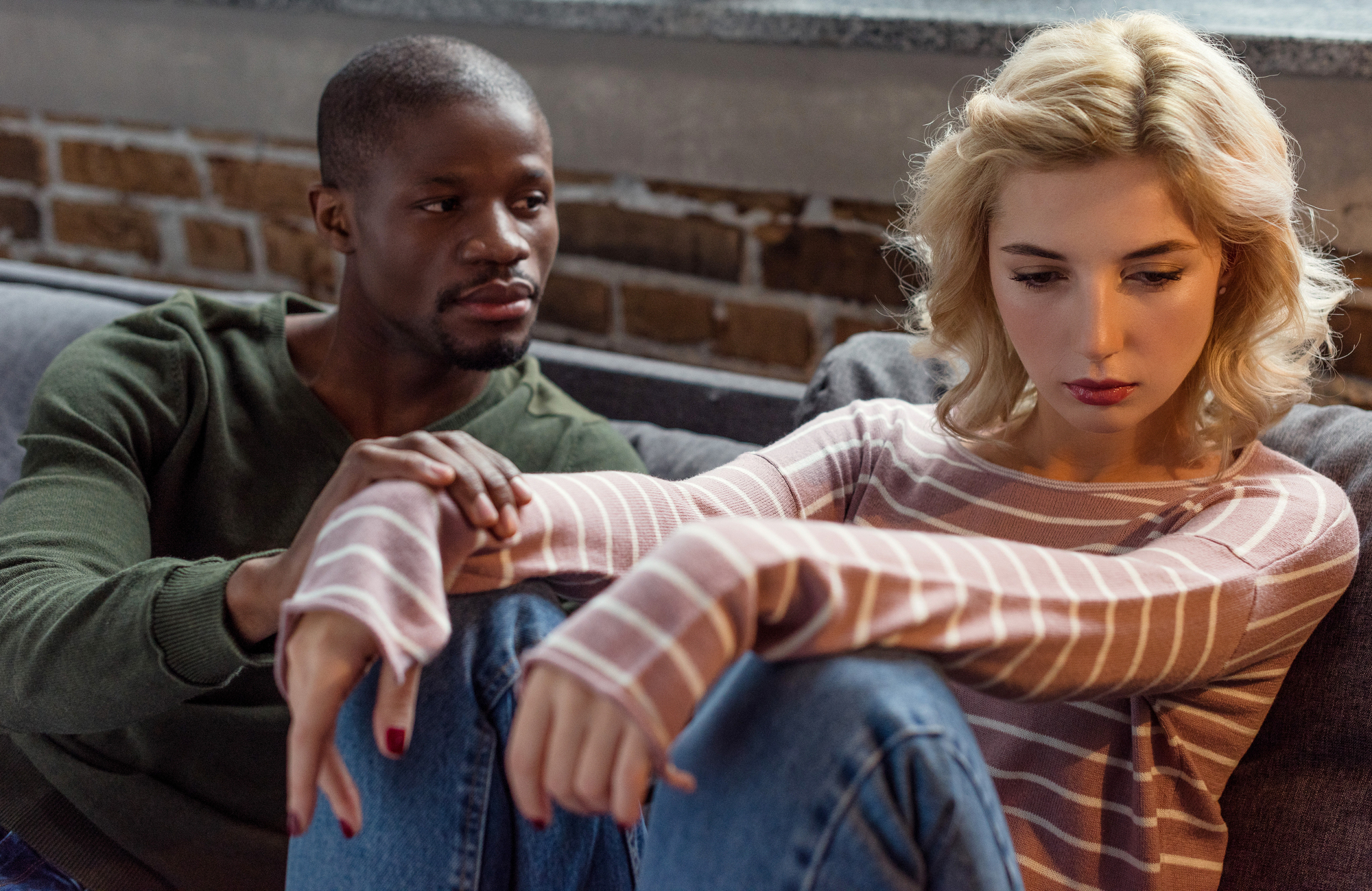 A man in a green sweater sits on a couch, gently touching the shoulder of a woman in a striped pink-and-white long-sleeve shirt and blue jeans. The woman looks down, appearing upset or deep in thought, while the man looks at her with concern.