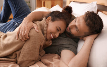 A couple lies on a bed in a cozy setting, with the woman smiling and nestling against the man. The man has his arm around her and is looking at her affectionately. Both appear relaxed and content, surrounded by warm, earthy tones of the bedding and room.