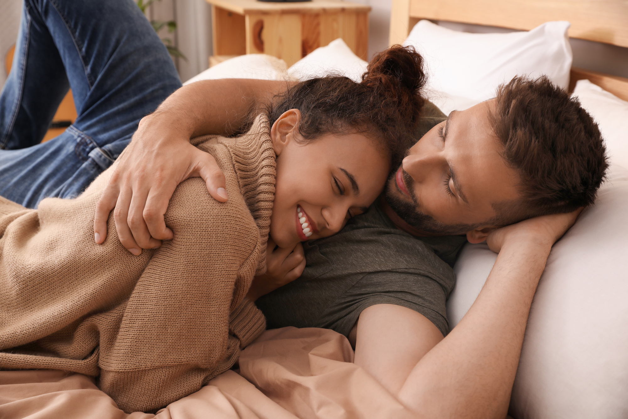 A couple lies on a bed in a cozy setting, with the woman smiling and nestling against the man. The man has his arm around her and is looking at her affectionately. Both appear relaxed and content, surrounded by warm, earthy tones of the bedding and room.