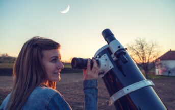 A woman with long hair and a nose piercing, wearing a denim jacket, looks through a large telescope during sunset. A crescent moon is visible in the sky. An out-of-focus house and trees are seen in the background.