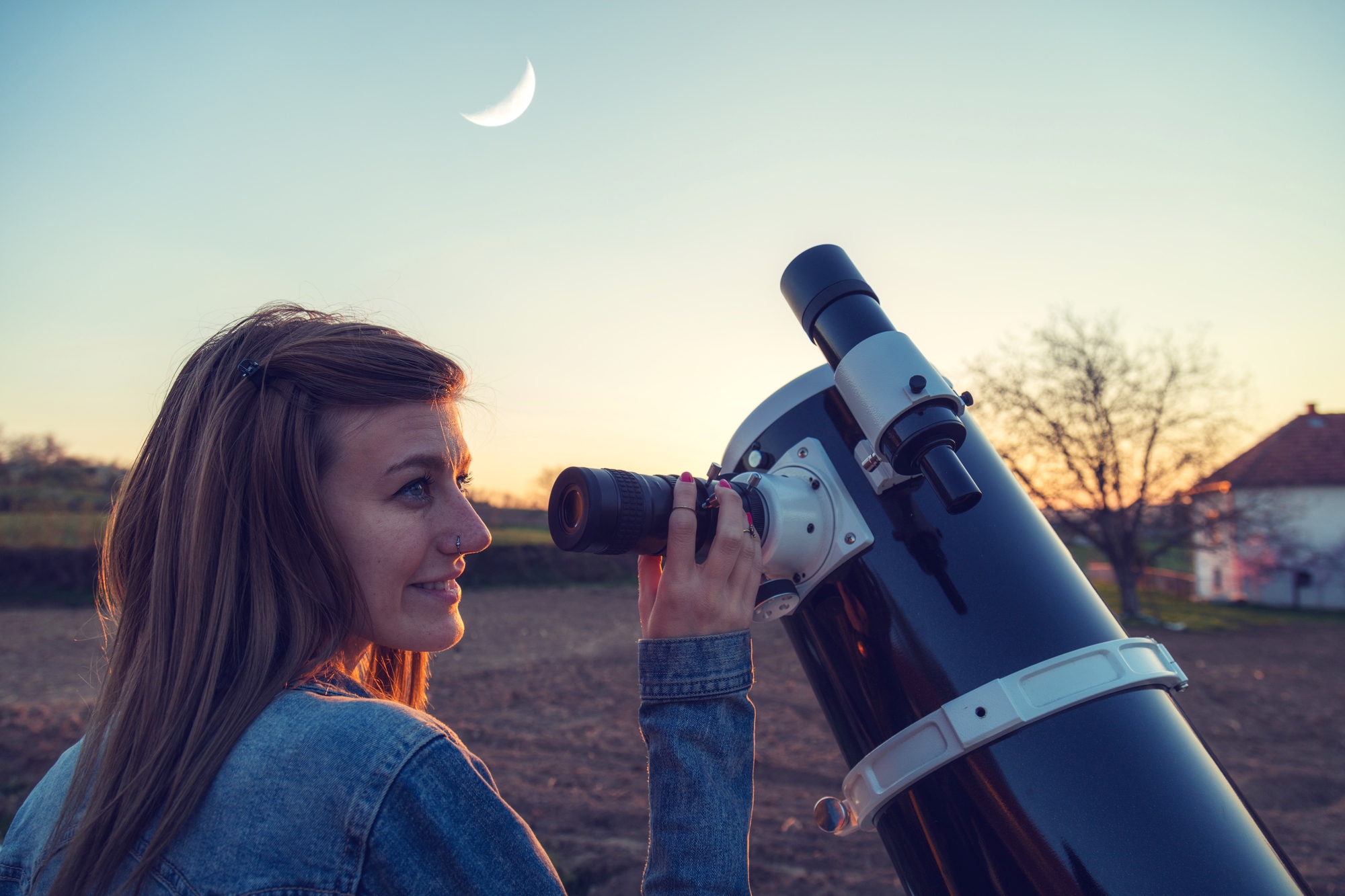 A woman with long hair and a nose piercing, wearing a denim jacket, looks through a large telescope during sunset. A crescent moon is visible in the sky. An out-of-focus house and trees are seen in the background.