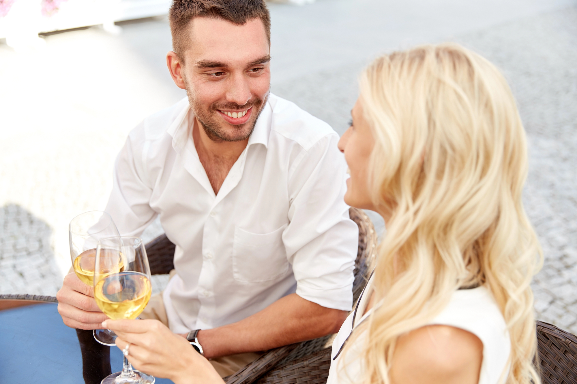 A man and woman sit at an outdoor table, smiling at each other while holding glasses of white wine. Both are wearing light-colored clothing, and the ambiance suggests a relaxed, pleasant conversation. The background is blurred but hints at a cobblestone surface.