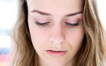 Close-up shot of a woman with light skin and wavy brown hair, looking down with closed eyes, displaying long eyelashes, and a neutral facial expression.