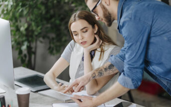 A man in a denim shirt and glasses points to a document on a table while a woman with long hair and a white vest listens attentively. They are working at a desk with a computer, notebook, and coffee cup in an office setting with greenery in the background.