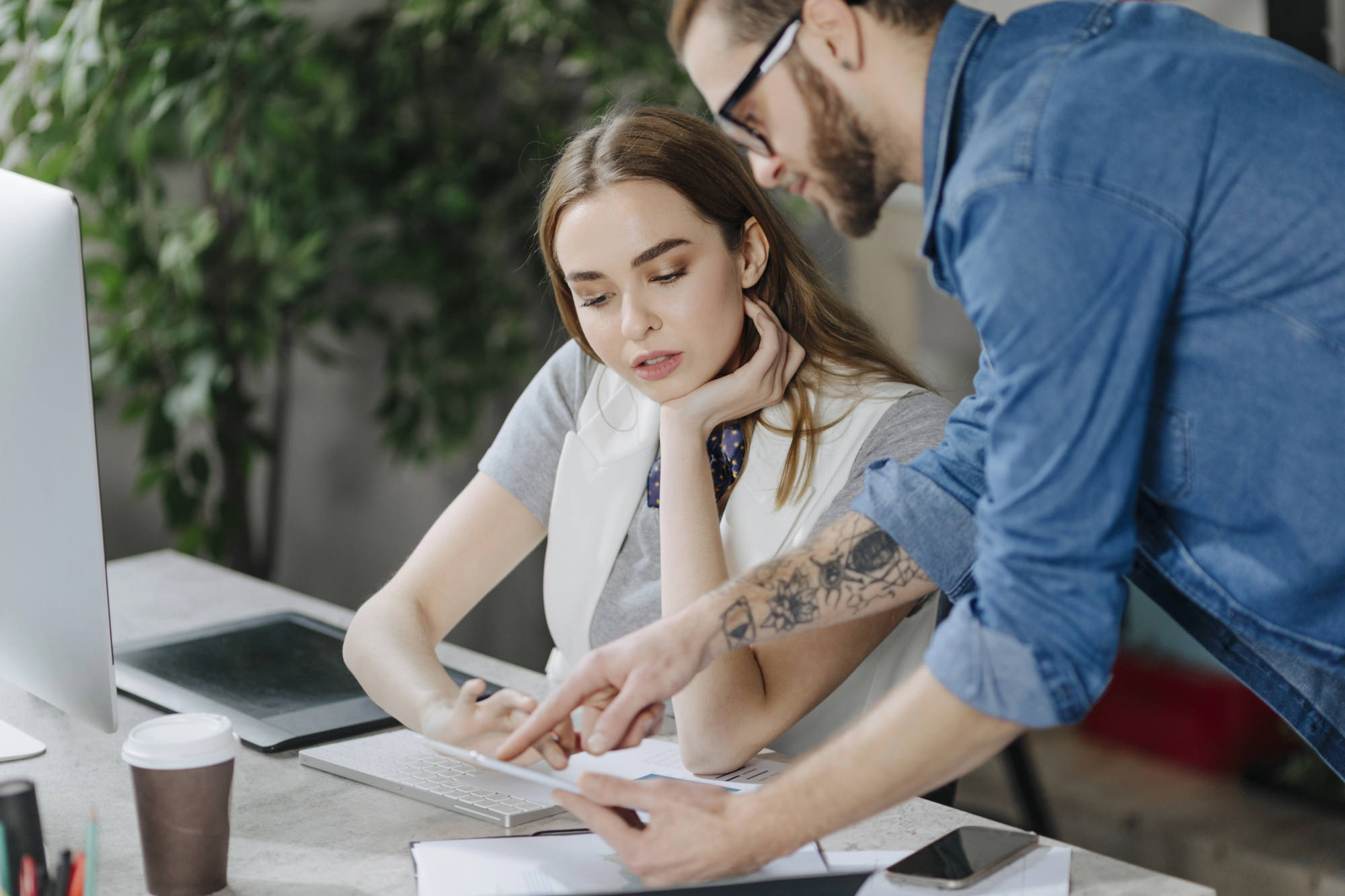 A man in a denim shirt and glasses points to a document on a table while a woman with long hair and a white vest listens attentively. They are working at a desk with a computer, notebook, and coffee cup in an office setting with greenery in the background.