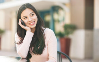 A young woman with long dark hair sits outdoors at a table, smiling and looking to her right while talking on a mobile phone. She is wearing a light pink sweater, and the background is softly blurred, suggesting an urban setting with buildings and greenery.