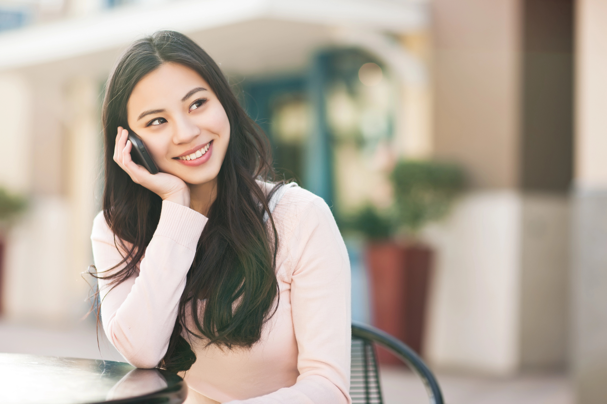 A young woman with long dark hair sits outdoors at a table, smiling and looking to her right while talking on a mobile phone. She is wearing a light pink sweater, and the background is softly blurred, suggesting an urban setting with buildings and greenery.