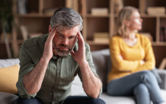 A distressed man with gray hair and beard holds his head in his hands while sitting on a couch. Behind him, a woman with blonde hair, wearing a yellow top and jeans, sits with her arms crossed, looking away. Shelves with books are in the background.