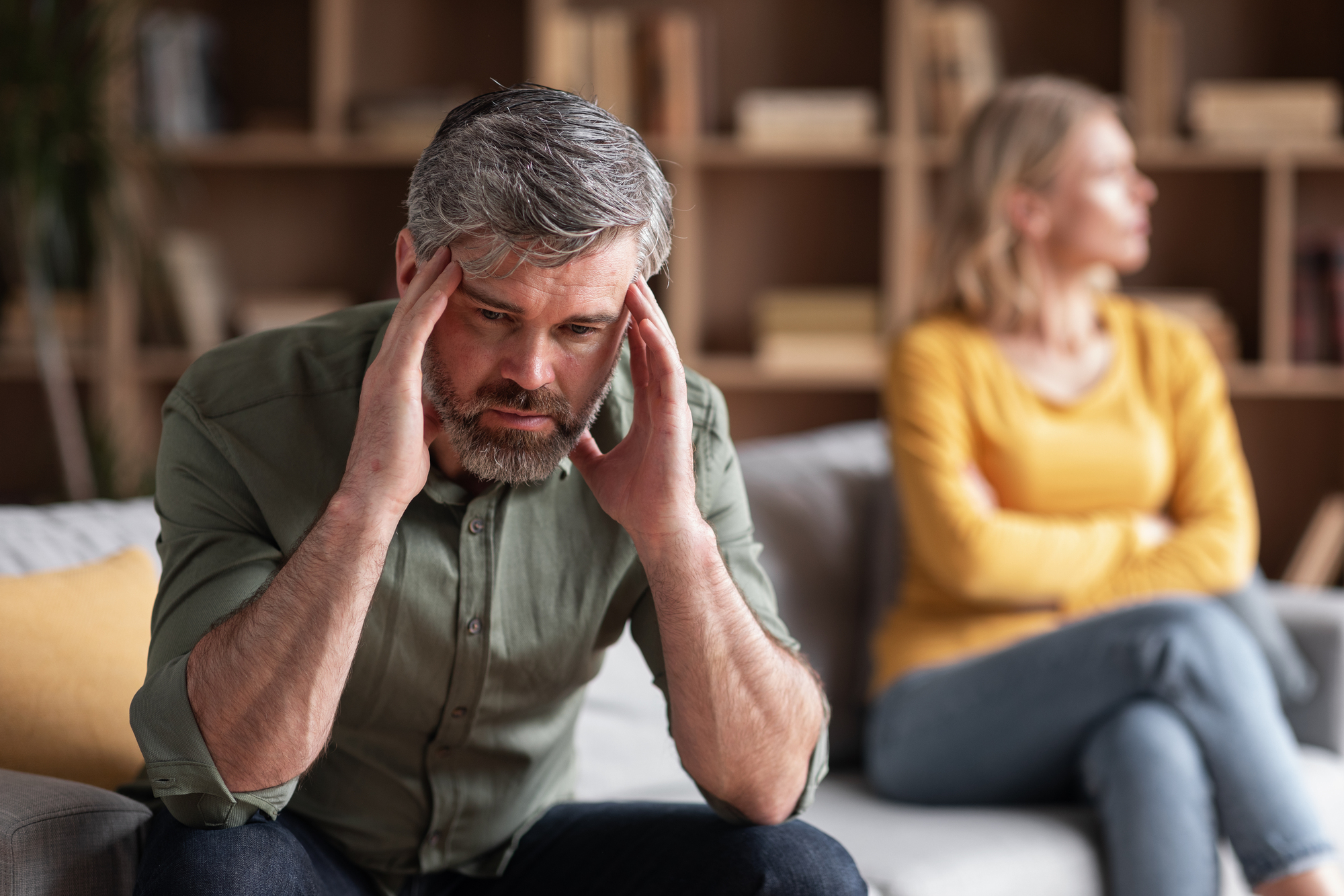 A distressed man with gray hair and beard holds his head in his hands while sitting on a couch. Behind him, a woman with blonde hair, wearing a yellow top and jeans, sits with her arms crossed, looking away. Shelves with books are in the background.