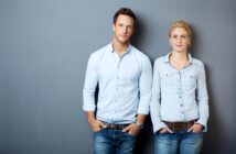 A man and a woman stand side by side against a gray background, both with hands in their pockets. They are wearing light blue button-up shirts and blue jeans. The man has short brown hair, and the woman has blonde hair pulled back. Both have a neutral expression.