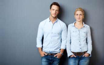 A man and a woman stand side by side against a gray background, both with hands in their pockets. They are wearing light blue button-up shirts and blue jeans. The man has short brown hair, and the woman has blonde hair pulled back. Both have a neutral expression.