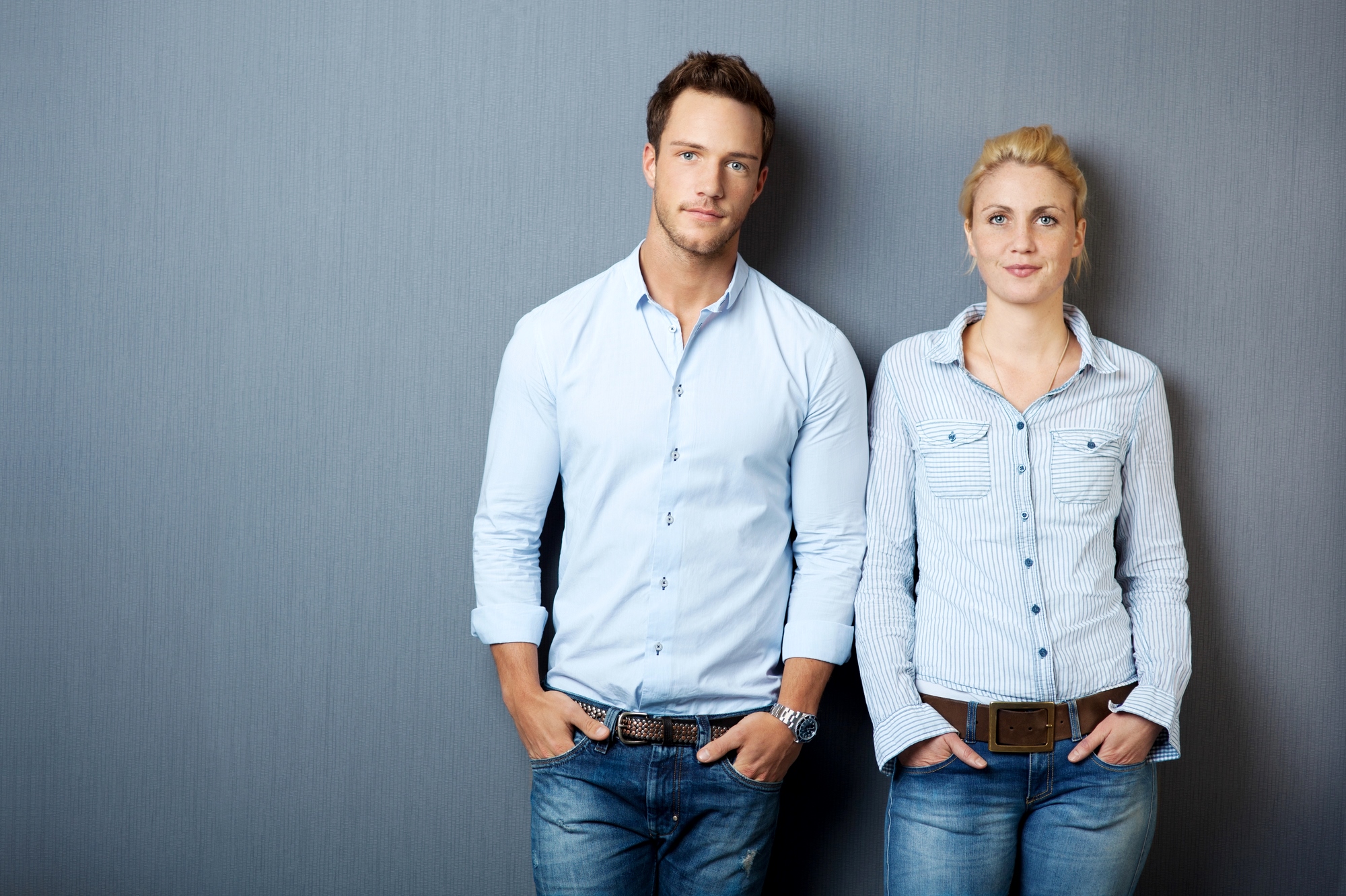 A man and a woman stand side by side against a gray background, both with hands in their pockets. They are wearing light blue button-up shirts and blue jeans. The man has short brown hair, and the woman has blonde hair pulled back. Both have a neutral expression.