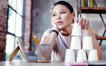 A woman sitting at a desk looks thoughtful and slightly bored as she rests her head on her hand. She is stacking white paper cups into a pyramid. The desk has pencils in a holder, a notepad, and a framed picture. The background includes shelves and a large window.