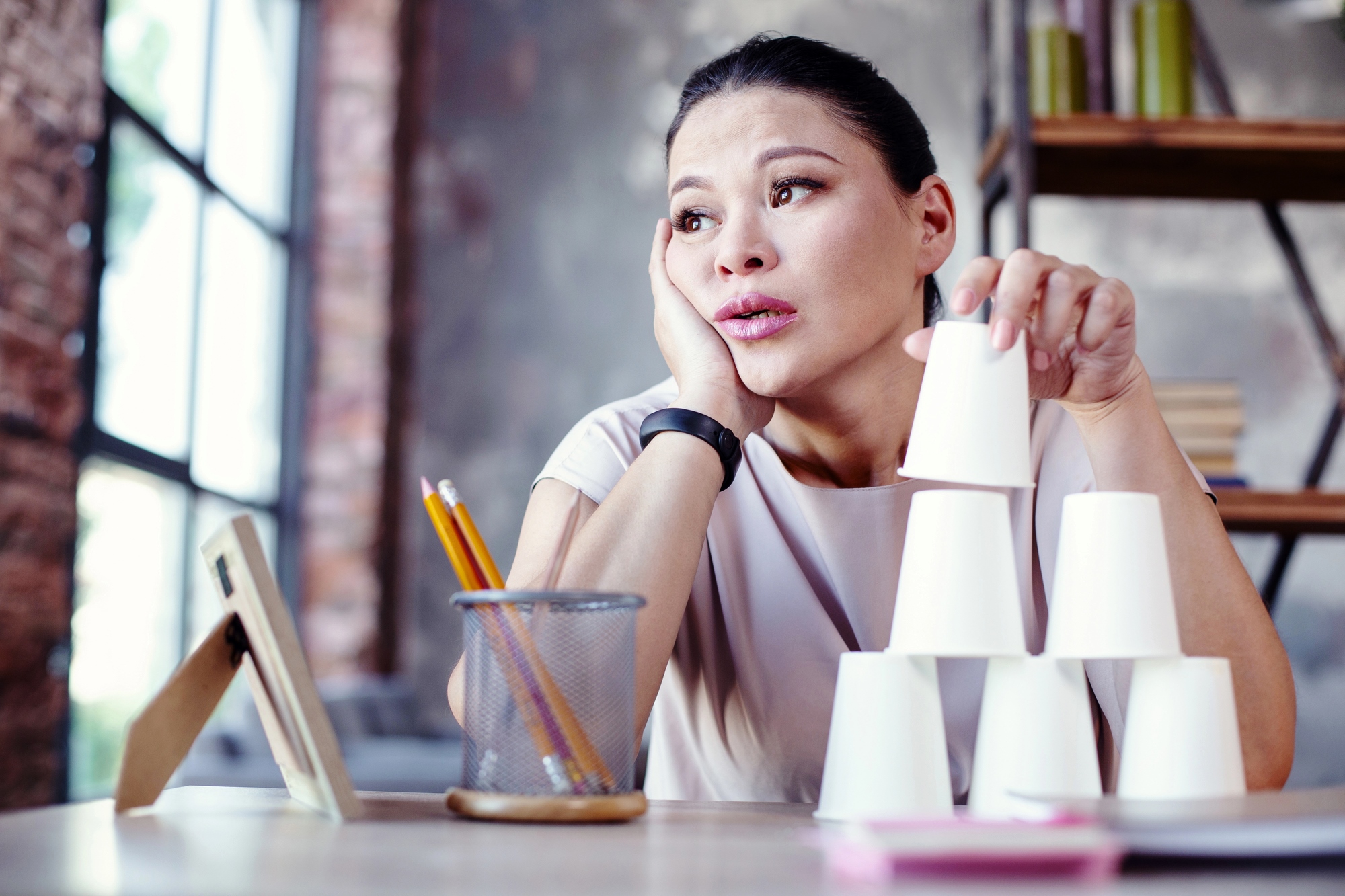 A woman sitting at a desk looks thoughtful and slightly bored as she rests her head on her hand. She is stacking white paper cups into a pyramid. The desk has pencils in a holder, a notepad, and a framed picture. The background includes shelves and a large window.