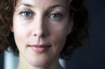 Close-up of a person with wavy brown hair and blue eyes, looking directly at the camera with a neutral expression. The background is dark and out of focus, highlighting the person's facial features.