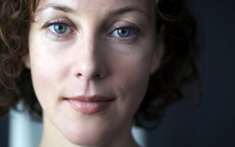 Close-up of a person with wavy brown hair and blue eyes, looking directly at the camera with a neutral expression. The background is dark and out of focus, highlighting the person's facial features.