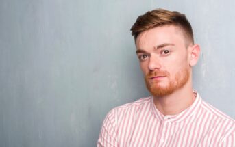 A young man with short brown hair and a neatly trimmed red beard stands against a light blue-gray background. He is wearing a pink and white striped shirt and looking directly at the camera with a neutral expression.