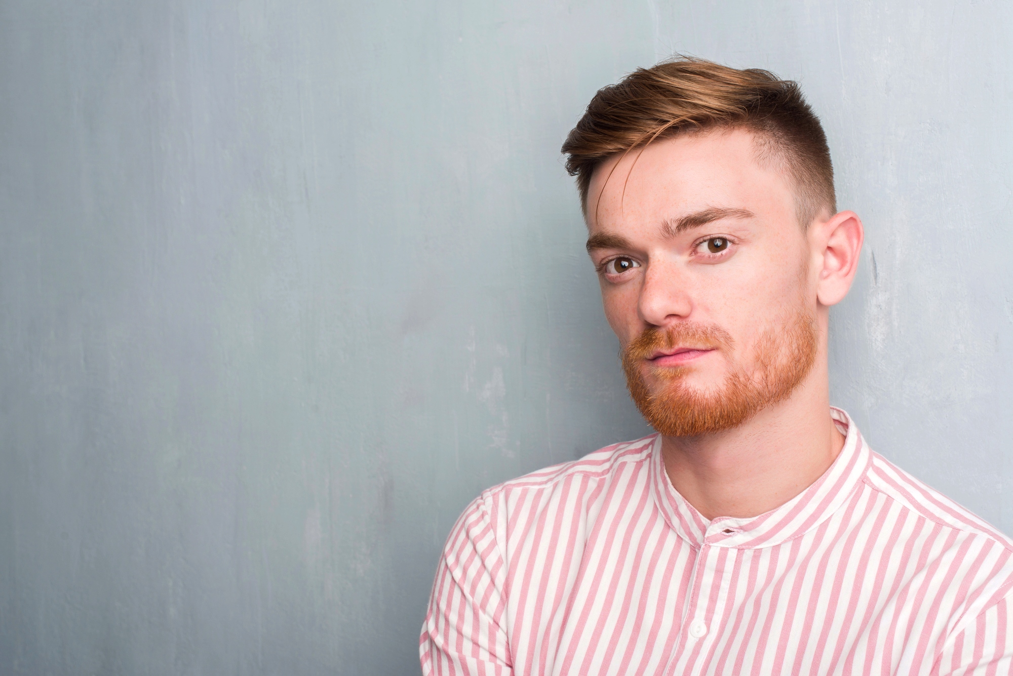 A young man with short brown hair and a neatly trimmed red beard stands against a light blue-gray background. He is wearing a pink and white striped shirt and looking directly at the camera with a neutral expression.