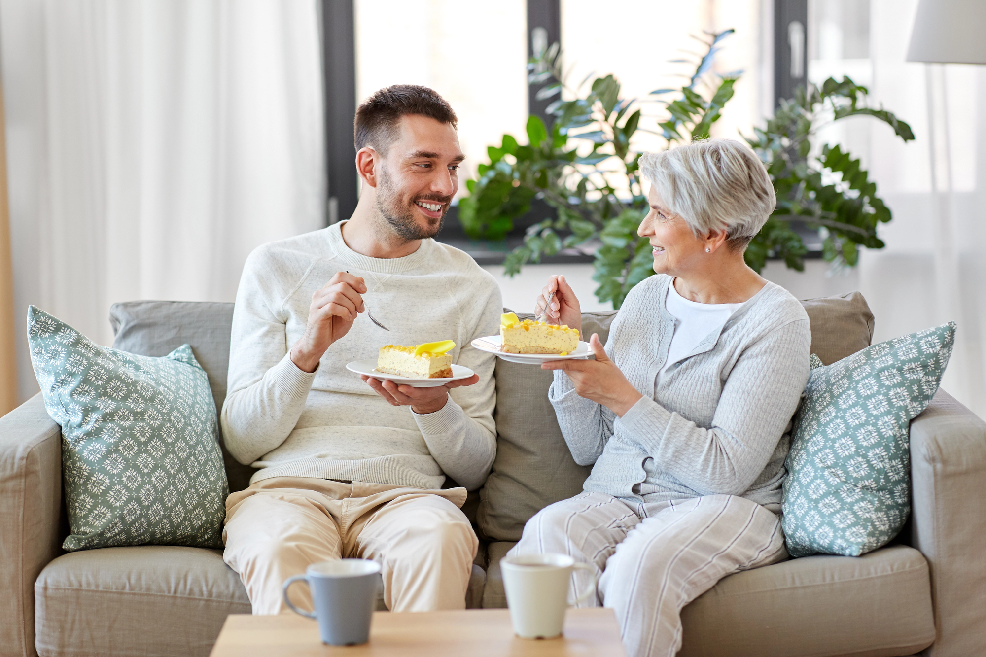 A man and an older woman sit on a couch in a bright, cozy living room, enjoying plates of cake together. They are smiling and engaging in conversation. Two mugs are on the table in front of them, and green cushions and plants add a touch of color to the scene.