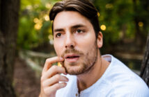 A man with medium-length brown hair and a beard, wearing a light-colored shirt, gazes thoughtfully while touching his lips with his finger. The background is a blurred, tree-filled outdoor setting.