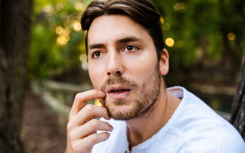 A man with medium-length brown hair and a beard, wearing a light-colored shirt, gazes thoughtfully while touching his lips with his finger. The background is a blurred, tree-filled outdoor setting.