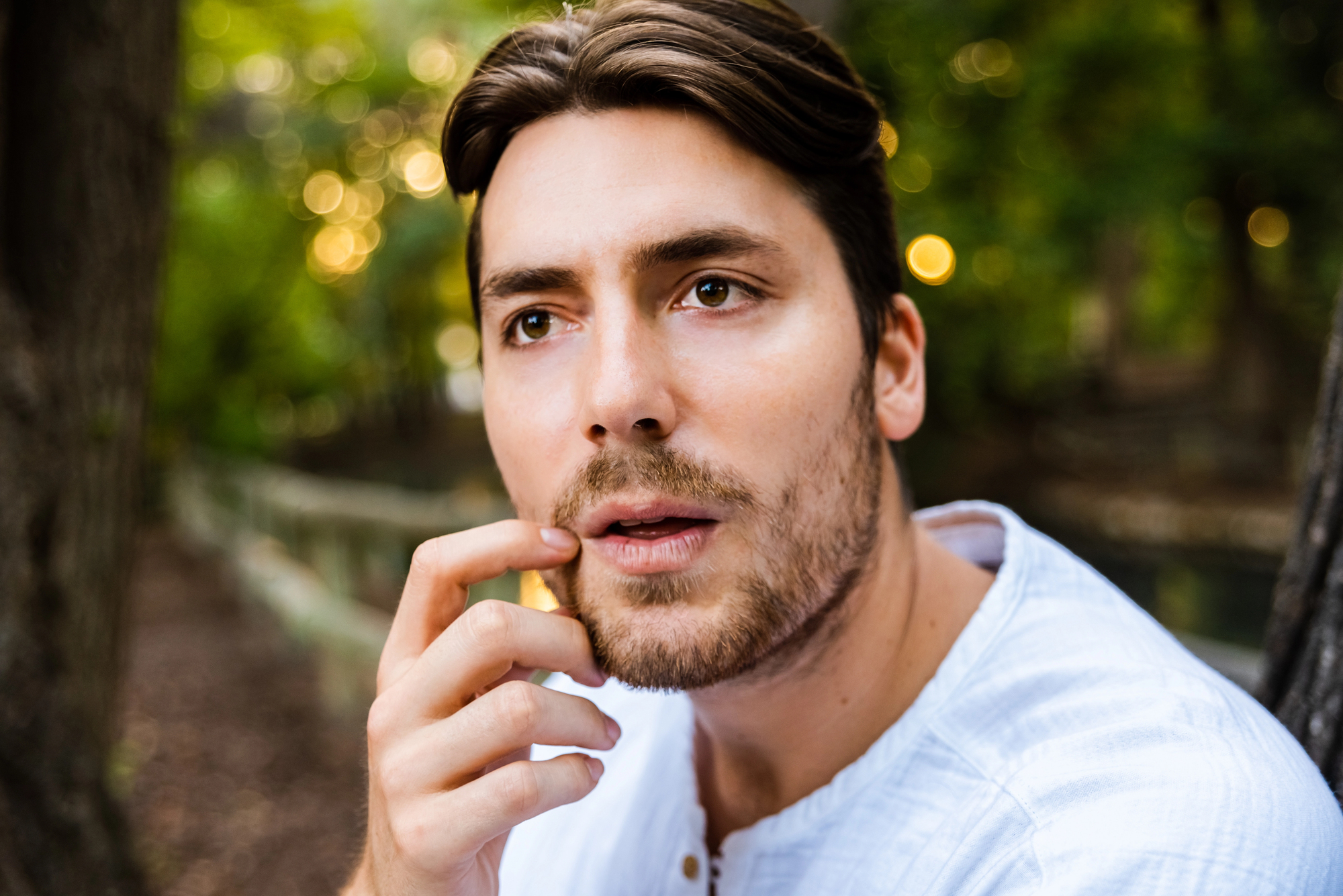 A man with medium-length brown hair and a beard, wearing a light-colored shirt, gazes thoughtfully while touching his lips with his finger. The background is a blurred, tree-filled outdoor setting.