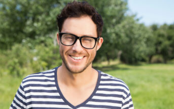 A man with short dark hair and glasses smiles while standing in a green outdoor setting. He is wearing a black and white striped V-neck shirt. The background is slightly blurred with lush trees and grass, suggesting a park or garden.