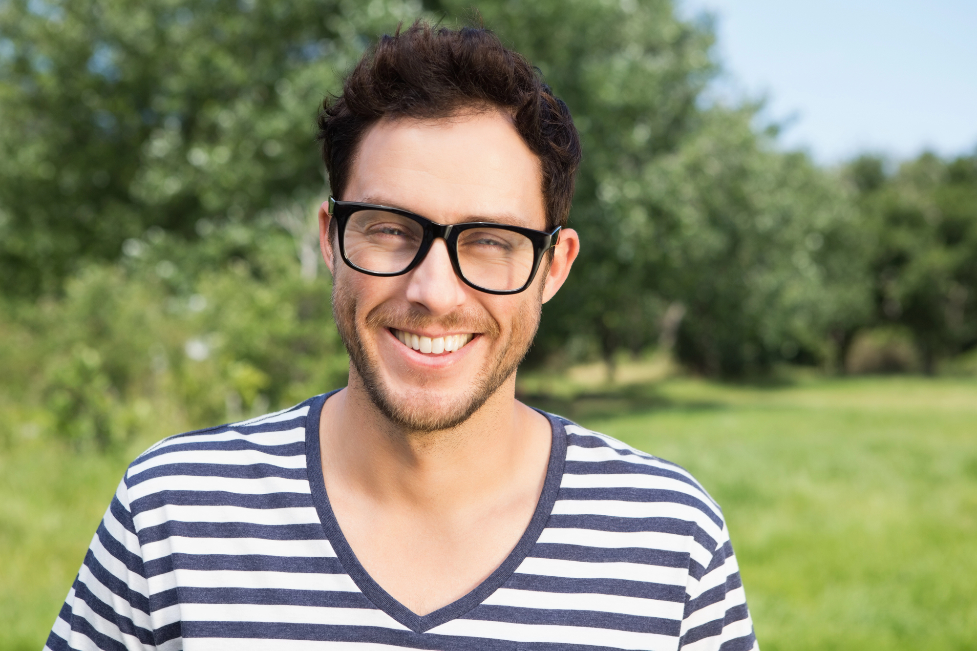 A man with short dark hair and glasses smiles while standing in a green outdoor setting. He is wearing a black and white striped V-neck shirt. The background is slightly blurred with lush trees and grass, suggesting a park or garden.