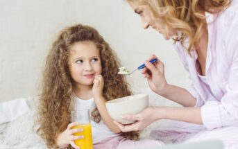 A young girl with curly hair holds a glass of orange juice and looks thoughtfully at an adult woman who is feeding her with a spoon from a bowl. They are sitting on a fluffy white surface, and both appear to be enjoying a casual moment together.