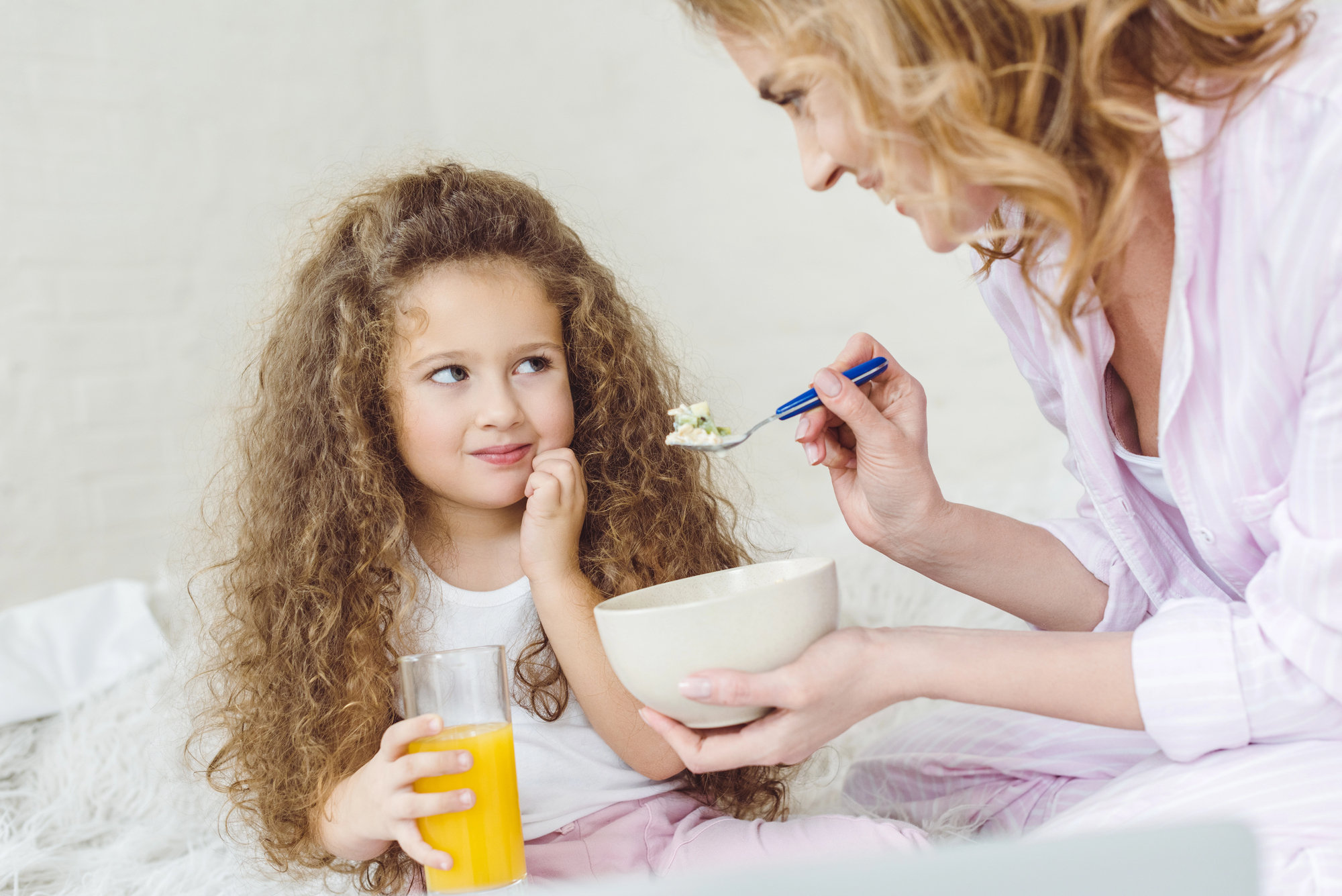 A young girl with curly hair holds a glass of orange juice and looks thoughtfully at an adult woman who is feeding her with a spoon from a bowl. They are sitting on a fluffy white surface, and both appear to be enjoying a casual moment together.