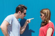 A man with dark hair and sunglasses is pointing a finger at a woman with long brown hair and sunglasses. The woman, dressed in a red top, stands facing the man in front of a corrugated blue wall, looking down with a neutral expression.