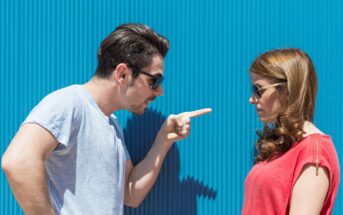 A man with dark hair and sunglasses is pointing a finger at a woman with long brown hair and sunglasses. The woman, dressed in a red top, stands facing the man in front of a corrugated blue wall, looking down with a neutral expression.