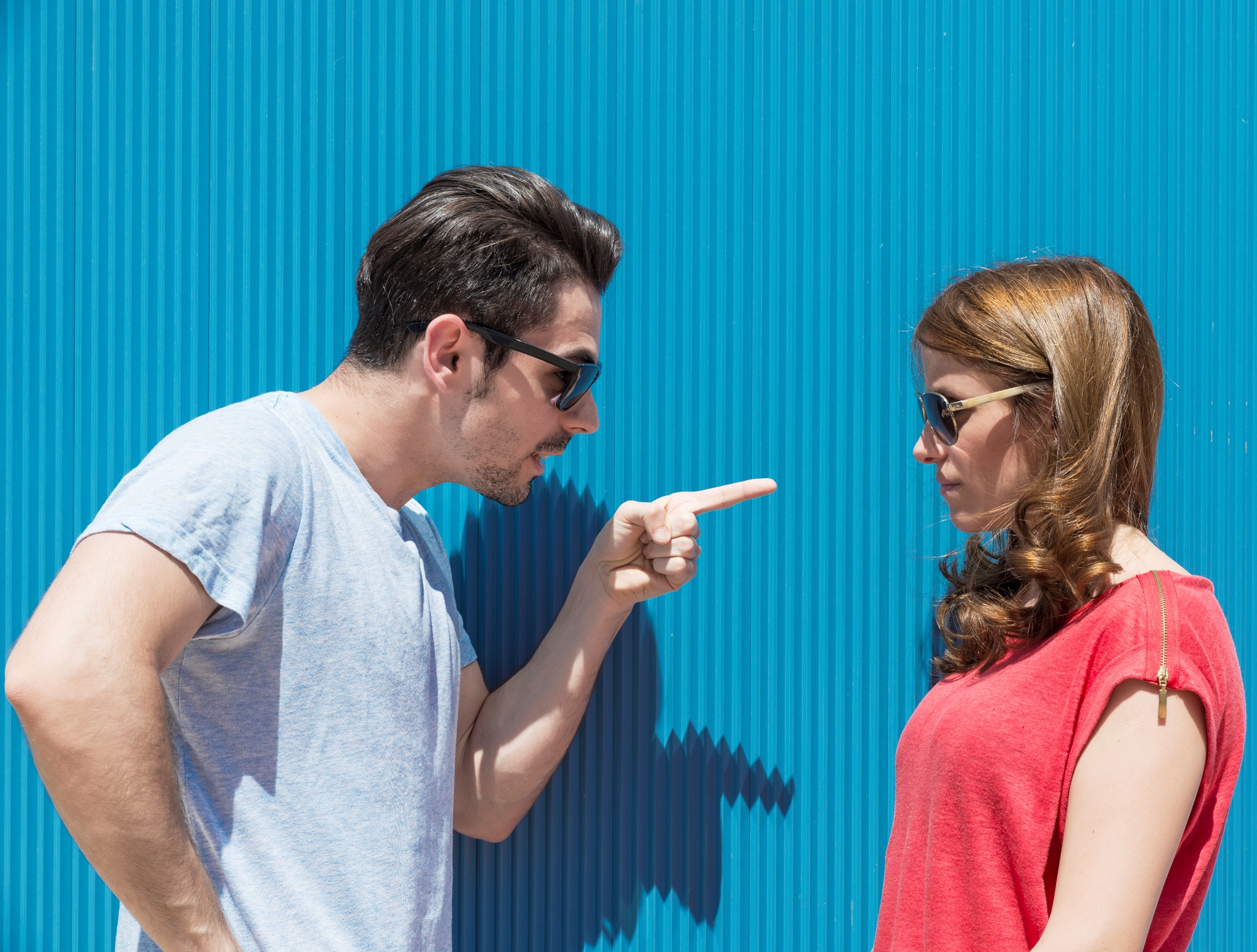 A man with dark hair and sunglasses is pointing a finger at a woman with long brown hair and sunglasses. The woman, dressed in a red top, stands facing the man in front of a corrugated blue wall, looking down with a neutral expression.