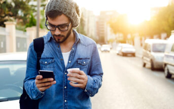 A man wearing sunglasses, a knitted hat, and a denim shirt holds a smartphone and a cup while walking down a sunlit street. He has a black backpack slung over one shoulder. There are cars parked along the side of the road and buildings in the background.