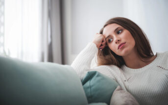 A woman with long brown hair, wearing a white sweater, rests her head on her hand while sitting on a couch. She looks thoughtfully into the distance with a pensive expression. Pillows in pastel colors are in the foreground.