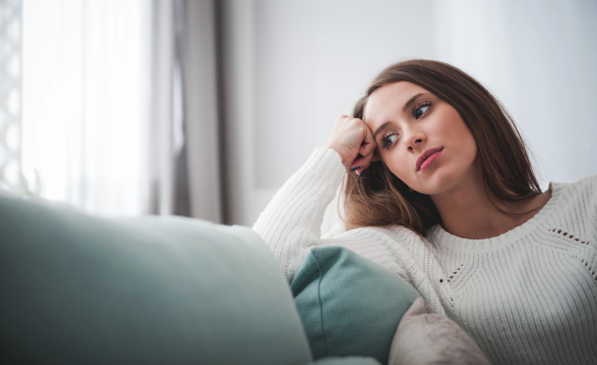 A woman with long brown hair, wearing a white sweater, rests her head on her hand while sitting on a couch. She looks thoughtfully into the distance with a pensive expression. Pillows in pastel colors are in the foreground.