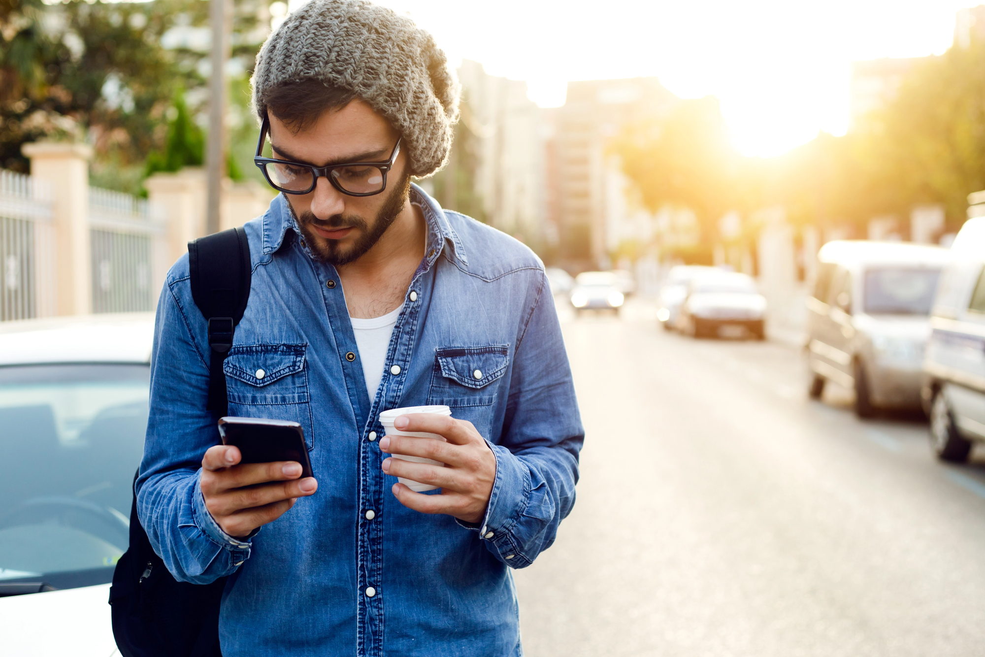 A man wearing sunglasses, a knitted hat, and a denim shirt holds a smartphone and a cup while walking down a sunlit street. He has a black backpack slung over one shoulder. There are cars parked along the side of the road and buildings in the background.